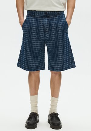 Személy tengerészkék kockás térd alatti rövidnadrágot, bézs zoknit és fekete bőrcipőt visel, kezét zsebében tartva áll.