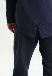 Giacca navy con una texture liscia, caratterizzata da un orlo spacco e una vestibilità rilassata, abbinata a pantaloni blu scuro.