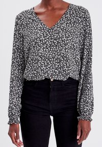 Schwarze Bluse mit langen Ärmeln und V-Ausschnitt, mit einem weißen Blumenmuster und gerüschten Bündchen. Kombiniert mit schwarzen, hoch geschnittenen Jeans.