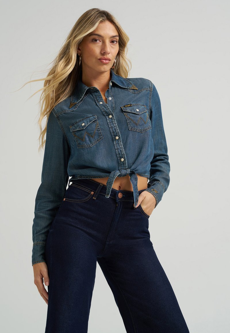 Denim shirt with two chest pockets, blue color, long sleeves, and a tied waist detail. Paired with high-waisted dark blue jeans.