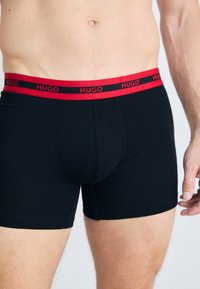 Männlicher Oberkörper in schwarzen Boxerbriefs mit einem roten Bündchen, auf dem der Markenname "HUGO" in roten Buchstaben steht.