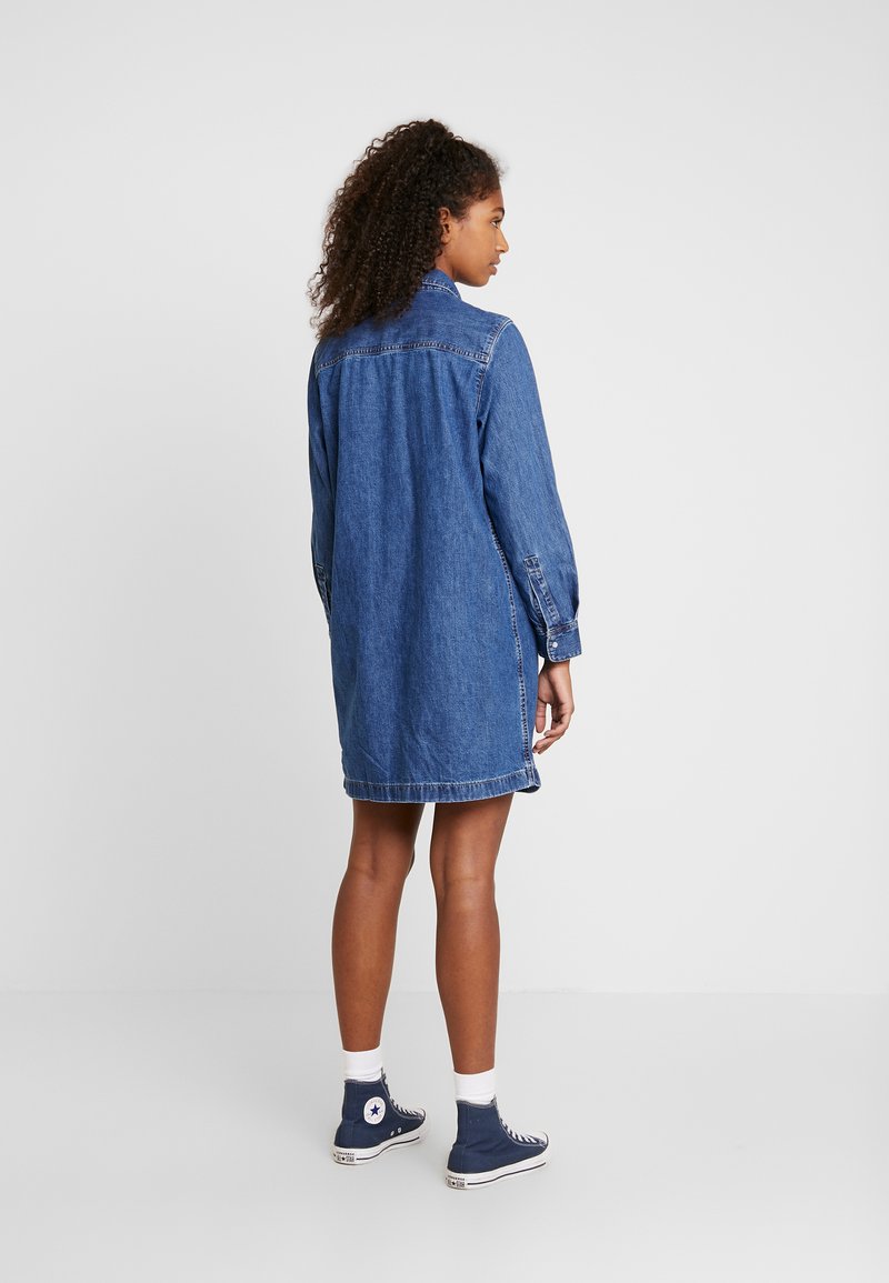 Robe chemise en denim bleu moyen, avec des manches longues, des poignets boutonnés et des poches latérales. Finition avec un ourlet arrondi et un détail de dos.
