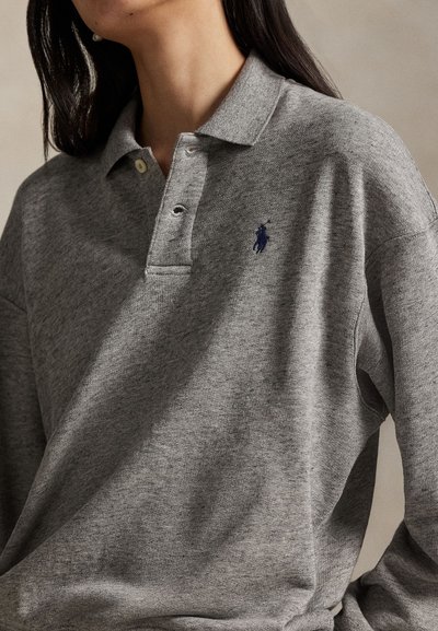 Nainen, joka käyttää harmaata pitkähihaista poolopaitaa, jossa on kaulus ja tummalla kirjailulla hevosen logo rinnassa.