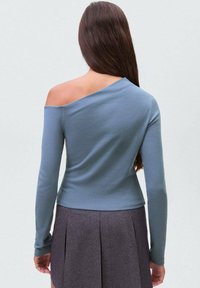 Dugmadna, off-shoulder bluza s dugim rukavima od mekanog materijala, koja ima uski kroj. Nosena s plisiranom suknjom koja ima glatku teksturu.