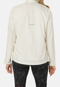 Vrouw in een licht beige lange mouwen jas en zwarte leggings met witte stippen, staande met haar rug naar voren.