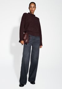 Pull en tricot bordeaux avec un subtil éclat, associé à un jean large gris foncé et des bottines noires. Sac à main en cuir marron avec cordon tenu à la main.