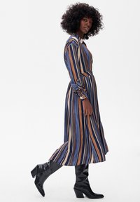 Sukienka z długim rękawem w paski w kolorach granatowym, beżowym i rdzy, z paskiem w talii i zapinanymi mankietami, połączona z wysokimi czarnymi botkami.