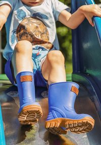 Blaue Gummistiefel mit braunen Klettverschlüssen werden von einem Kind getragen, das eine Metallrutsche hinunterrutscht. Die Sohle ist strukturiert und die Farben sind lebhaft.