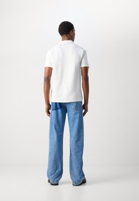 Weißes Poloshirt mit kurzen Ärmeln aus strukturiertem Stoff, kombiniert mit hellblauen weiten Jeans mit einer aufgesetzten Tasche. Schwarze Schuhe runden das Outfit ab.