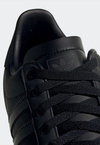 Gros plan sur une chaussure de sport Adidas en cuir noir avec des lacets noirs texturés et le logo tréflé embossé d'Adidas sur la languette.