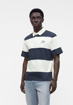 Homme portant un polo à manches courtes rayé bleu marine et blanc avec un logo sur la poitrine et un jean bleu clair, debout devant un fond uni.