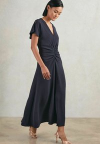 Navy-maxikleid mit V-Ausschnitt, kurzen Ärmeln und gerafftem Vorderteil. Glattes Material und fließende Silhouette. Kombiniert mit metallischen Riemchensandalen.