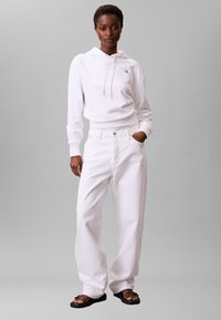 Sudadera blanca de algodón con logo y cordones, combinada con vaqueros blancos de tiro alto y pierna ancha; la modelo lleva sandalias marrones con tiras.