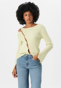 Maglione a coste di un leggero giallo, con design off-shoulder, bottoni decorativi lungo il lato e maniche lunghe abbinate a jeans blu.