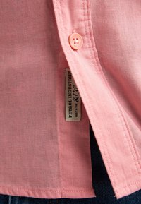 Gros plan de l'ourlet d'une chemise rose boutonnée avec une étiquette en tissu « Petrol Industries & Co » et une partie d'un jean bleu foncé visible en dessous.