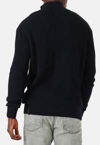 Mann in einem marineblauen Rippstrickpullover und hellgrauen Jeans, von hinten vor einem schlichten weißen Hintergrund gezeigt.