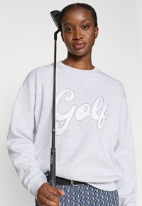 Sweatshirt cinza com a palavra "Golf" em letras bordadas, usado com calças azuis e brancas estampadas. Um taco de golfe é segurado em uma das mãos.