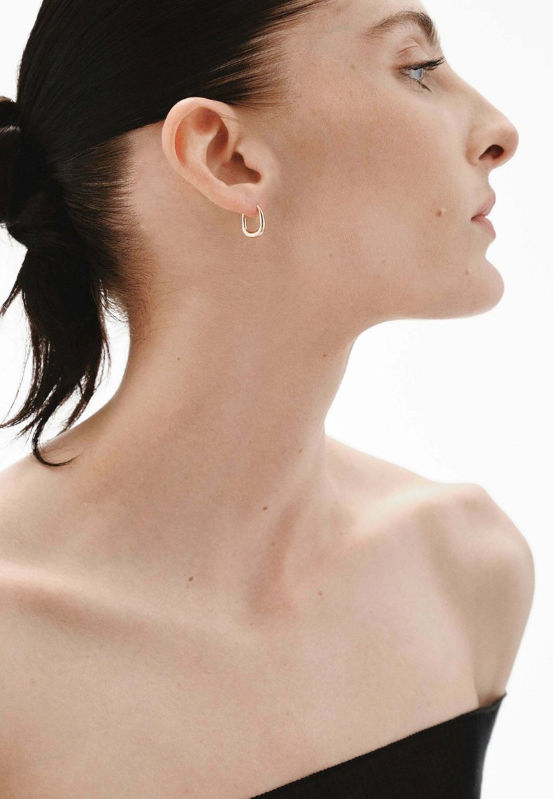 Pendiente de aro dorado con un diseño en forma de U. El modelo tiene el cabello lacio y lleva una blusa negra, lo que resalta el pendiente de manera destacada.