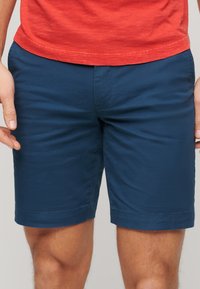 Shorts bleu marine, arrivant aux genoux, fabriqués dans un tissu lisse avec un devant plat, une coupe droite et des poches latérales. Pas de motifs ni de pièces métalliques visibles.