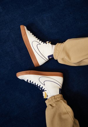 Weiße High-Top-Sneaker mit braunen Sohlen und marineblauen Schnürsenkeln, getragen zu beigen Jogginghosen vor einem dunkelblauen Teppichhintergrund.