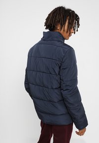 Marineblå pufferjakke med høj krave, quiltede sektioner og en glat tekstur. Har en tætsiddende pasform og minimalistisk design.