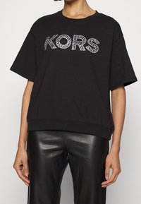 Persona vistiendo una camiseta negra de gran tamaño con "KORS" en piedras preciosas plateadas y pantalones de cuero negros, mostrada de cuello a caderas.