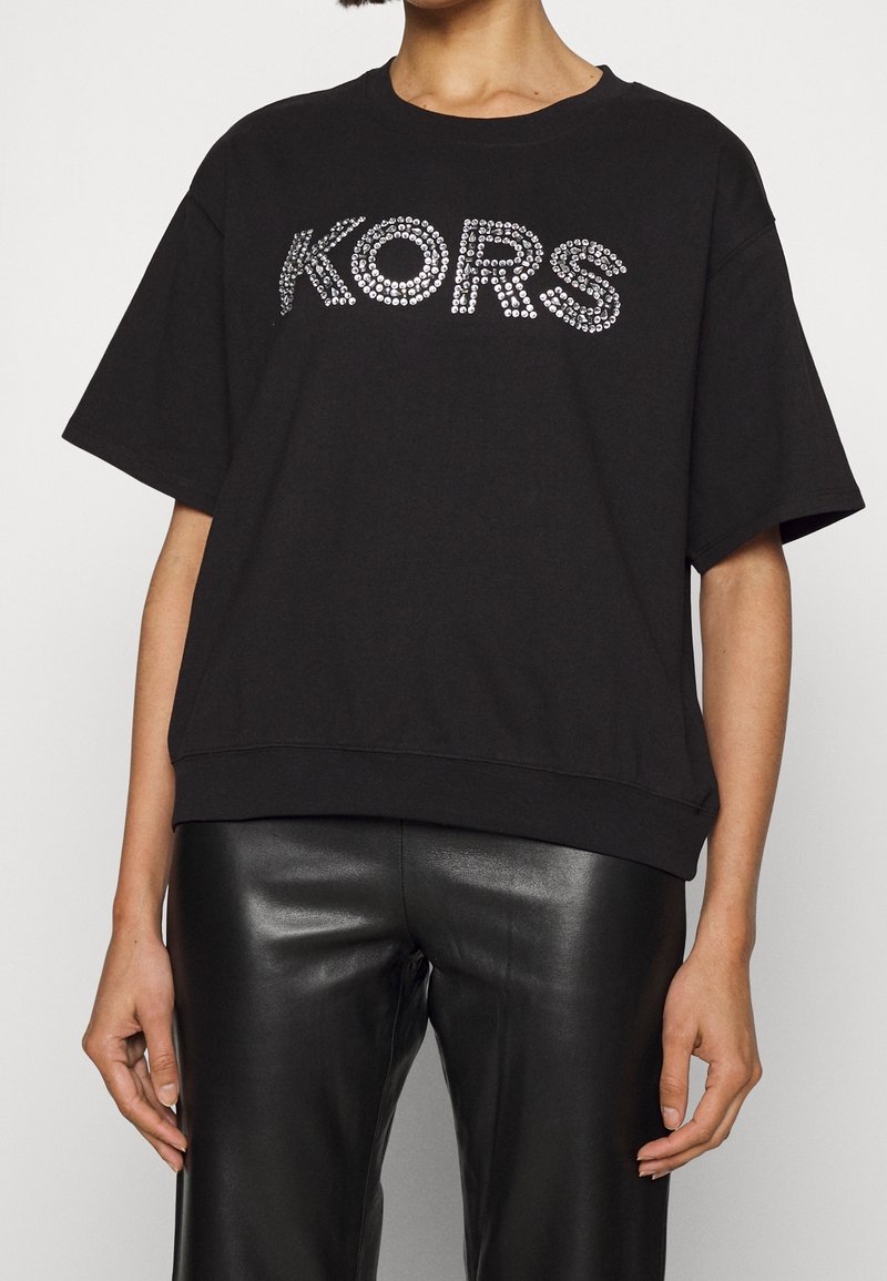 Persona vistiendo una camiseta negra de gran tamaño con "KORS" en piedras preciosas plateadas y pantalones de cuero negros, mostrada de cuello a caderas.