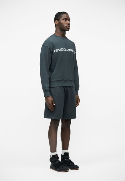 Jeune homme portant un sweat-shirt foncé avec le texte « ENDORPHIN », un short assorti, des chaussettes blanches et des baskets montantes noires, debout devant un fond uni.