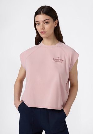 Femme aux longs cheveux foncés portant un débardeur rose Champion Training Club et un pantalon foncé, les mains dans les poches, expression neutre.