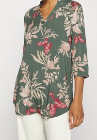 Grünbluse mit floralem Muster und drei Viertelärmeln, mit Schattierungen von rosa und roten Blumen sowie Schmetterlingen und einem V-Ausschnitt.