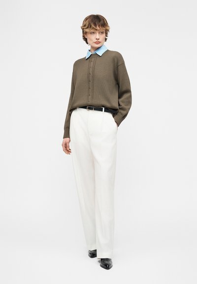 Νέος άνθρωπος στέκεται φορώντας λευκό παντελόνι ψηλόμεσο, χακί πουλόβερ πάνω από μπλε πουκάμισο με γιακά, μαύρη ζώνη και μυτερά μαύρα παπούτσια σε λευκό φόντο.