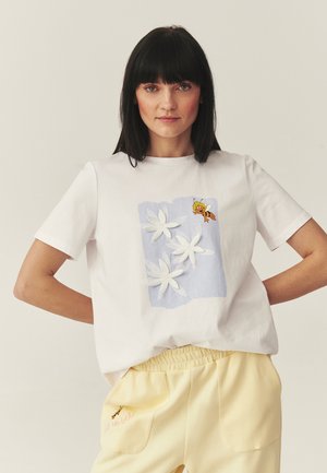 Kobieta w białym t-shircie z trzema białymi kwiatami i kreskówkową pszczołą, w połączeniu z jasnżółtymi spodniami z haftem pszczoły na kieszeni.