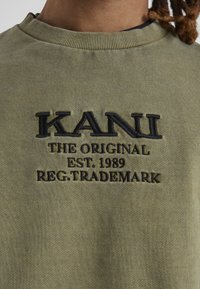 Olivgrön sweatshirt med ribbat runt halsen, med svart broderad "KANI" logotyp och texten "THE ORIGINAL EST. 1989 REG.TRADEMARK."