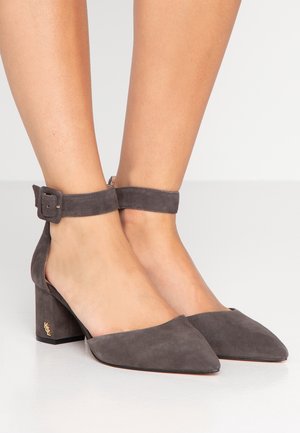 Voeten met grijze suède pumps met een blokhak, spitse neus, enkelbandjes en een klein gouden logo op de hak.