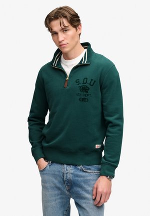 Junger Mann trägt einen dunkelgrünen Quarter-Zip-Pullover mit dem Text "S.O.U ATH. DEPT.", hellblaue Jeans und ein weißes T-Shirt darunter.