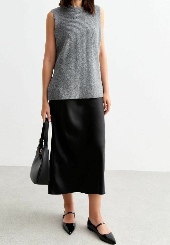 Femme portant un haut tricoté gris sans manches, une jupe midi noire, des ballerines pointues noires, et tenant un sac à main noir à son côté.