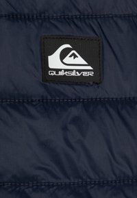 Námornícka modrá prešívaná tkanina s horizontálnymi švami. Obsahuje čiernu obdĺžnikovú etiketu s bielym logom a textom "Quiksilver" uprostred.