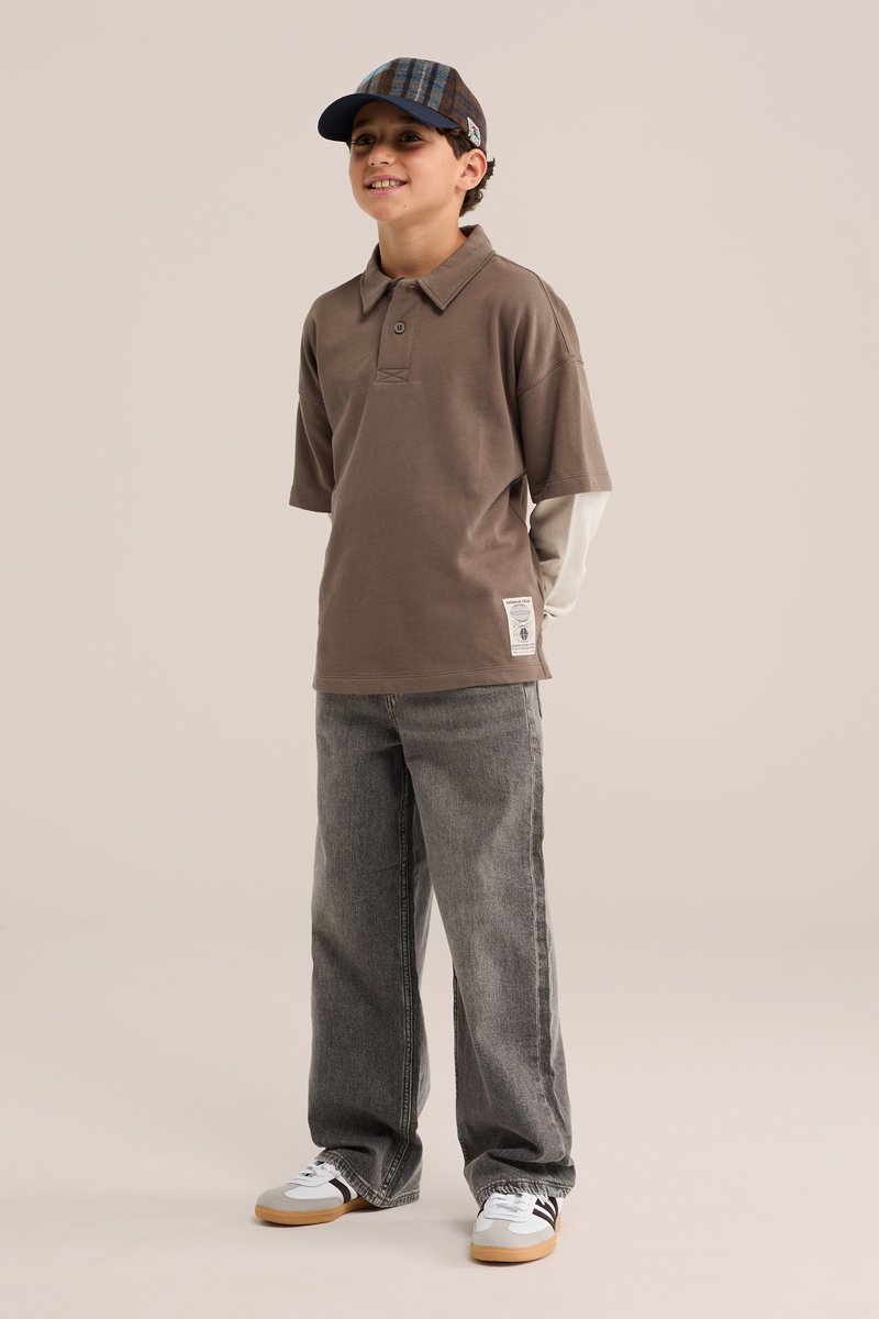 Garçon portant un polo marron à manches courtes par-dessus une chemise à manches longues, jean gris, baskets blanches et une casquette à carreaux, debout et souriant.