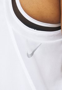 Camiseta deportiva blanca sin mangas con una tela texturizada, que presenta una capa de malla negra debajo y un logo plateado de Nike en la parte frontal.
