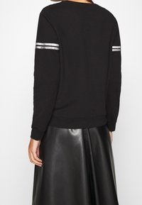 Donna che indossa un top a maniche lunghe nero con due strisce argento su ciascuna manica e una gonna in pelle nera, vista da dietro.