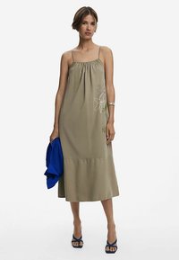 Khakifärgad maxiklänning med samlad halsringning och blommig broderi. Har tunna band och en flerstegs fåll. Matchas med en blå jacka.