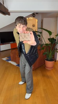 Rudas kortelių laikiklis su raštuota tekstūra, kuriame yra trikampio formos logotipas; laikomas asmens, dėvinčio paminkštintą liemenę ir pilkus kelnes, fone matomi vidaus augalai.