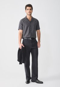 Camisa de manga corta en gris oscuro con botones y un bolsillo, combinada con pantalones negros. El hombre sostiene un suéter negro y lleva botas negras.