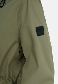 Olivgrüne Jacke aus strukturiertem Stoff mit einem schwarzen Patch am Ärmel. Mit Druckknöpfen zum Schließen und einem lässigen Schnitt.