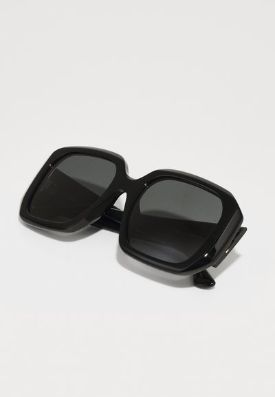Occhiali da sole oversized neri con un design geometrico massiccio, lenti quadrate, finitura lucida e dettagli laterali per un supporto aggiuntivo.