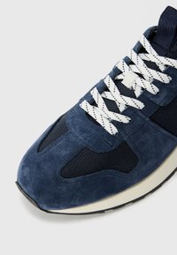 Tumesinine nubukist sneaker, millel on võrgupaneelid, valged nööbid ja kreemjas vahekolle. Ümar nina koos tekstuuriga pinnaga ja must tald.