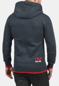 Dark blue hoodie with a red accent at the hem. Features a large hood and embroidered text near the bottom: "! SLD Since 1993."