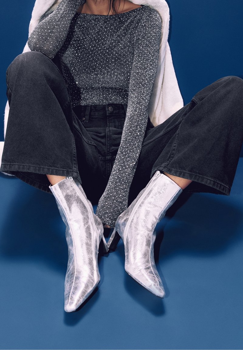 Personne assise les jambes écartées, portant un haut à manches longues gris pailleté, un pantalon large noir et des bottes à talons hauts pointues argentées brillantes sur un fond bleu.