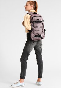 Zaino viola texturizzato con spallacci regolabili, tasche in rete e fibbie nere. Indossato sulla schiena di una persona con jeans grigi e scarpe chiare.