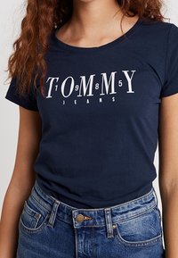 Marinblå bomull T-shirt med vit "TOMMY JEANS" tryck med numeriska accenter. Slim fit, korta ärmar och rund halsringning.